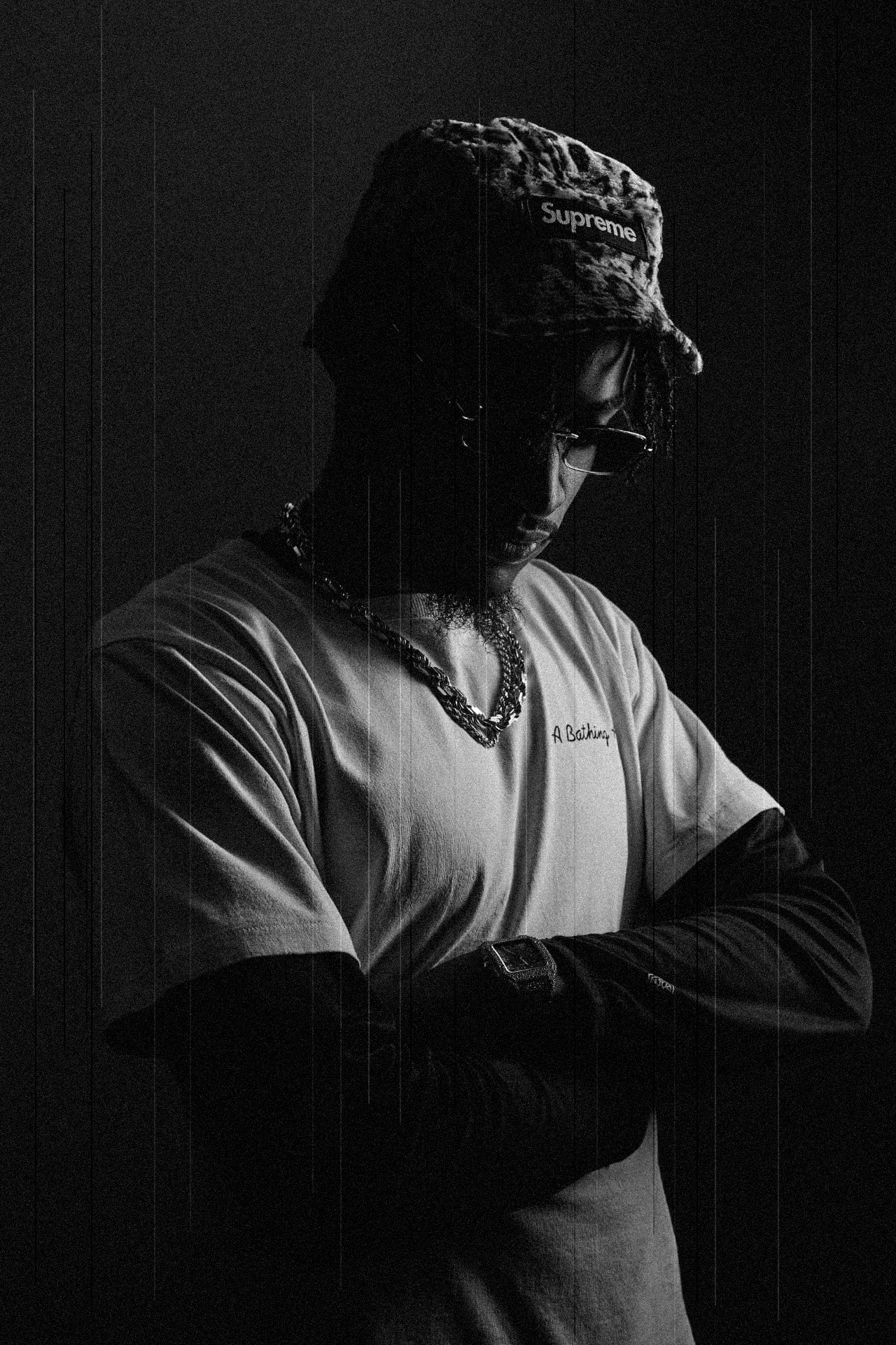 Touché Noir in low-key light with arms crossed, printed cap and dark sunglasses