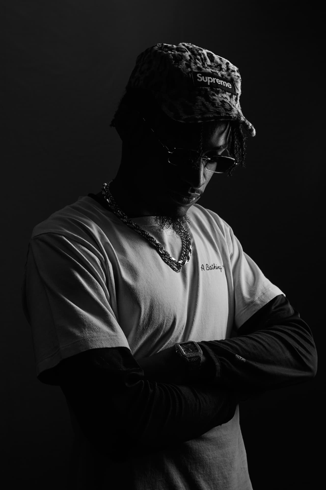 Touché Noir in low light, arms crossed, head lowered beneath the brim of his cap